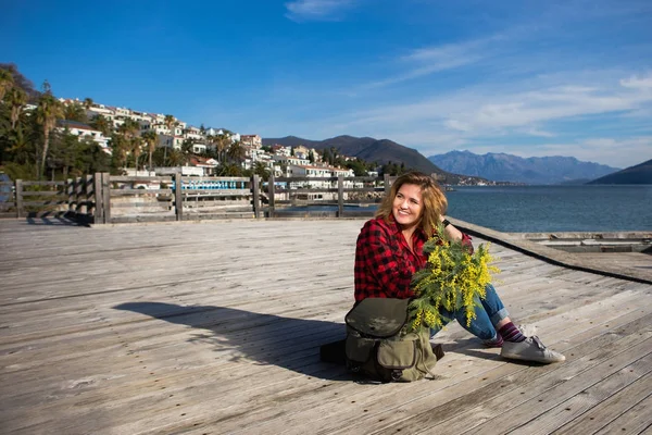 Счастливая девушка путешественник сидит на деревянном пирсе и держит букет цветов мимозы в руках . — стоковое фото