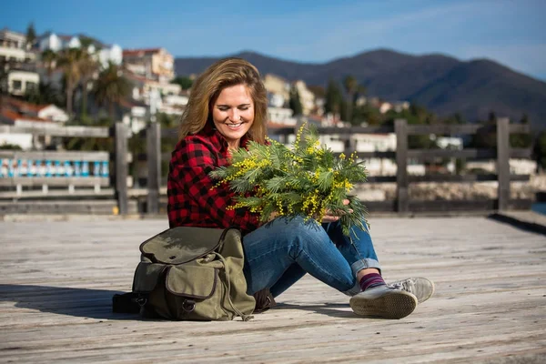 Glückliche Reisende sitzt auf einem Holzsteg und hält einen Strauß Blumen von Mimosen in den Händen. — Stockfoto