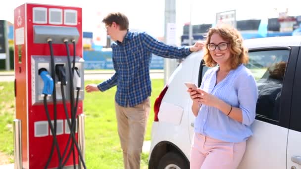 在电动车充电站的年轻夫妇微笑 男子收费汽车和妇女站在智能手机 — 图库视频影像