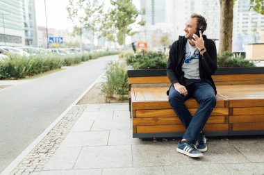 Neşeli adam şehirde bir bankta oturur ve telefonda konuşur.