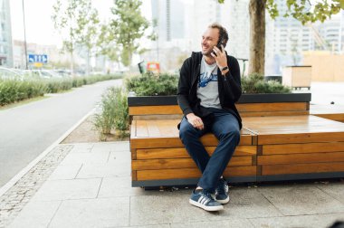 Neşeli adam şehirde bir bankta oturur ve telefonda konuşur.