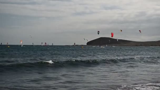 Veel kleurrijke vliegers op het strand en kitesurfers paardrijden golven tijdens winderige dag — Stockvideo