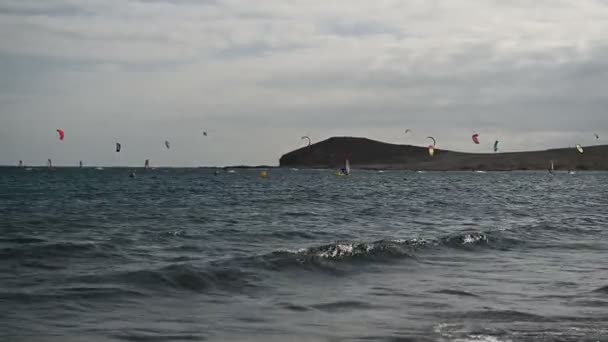 Många färgglada drakar på stranden och kitesurfare rider vågor under blåsig dag — Stockvideo