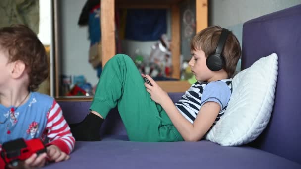 Un adolescente juega en el teléfono con auriculares, y el hermano menor juega un juguete - un coche — Vídeo de stock