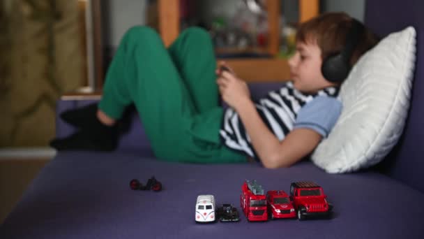 Een tiener speelt op de telefoon met een koptelefoon, en de jongere broer speelt een speeltje - een auto — Stockvideo