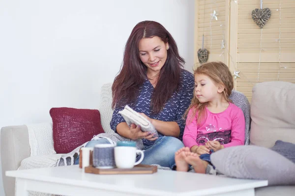 Maman et sa fille sont assises sur le canapé à la maison et lisent un livre. Joyeux famille aimante. Mère et sa fille enfant fille jouent. Bon temps à la maison . — Photo