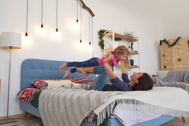 Anne ve kızı evde, yatakta oynuyorlar. Anne sırt üstü yatıyor ve kızını ayaklarının üzerinde tutuyor. Kadın gülerken kız mutlu görünüyor.