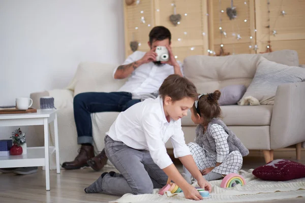 Papa et les enfants s'amusent ensemble à la maison, papa s'assoit sur le canapé et photographie ses enfants. fils et fille jouent sur le sol — Photo