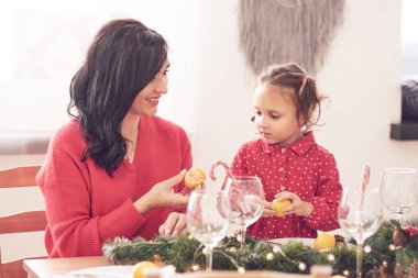 Neşeli anne ve kızı gülüyor, Noel ağacıyla eğleniyorlar ve mutfak masasında yıldız şeklinde kurabiyeler yiyorlar. Kırmızı giysiler içinde ren geyiği boynuzu ve Noel Baba şapkası takıyorlar..