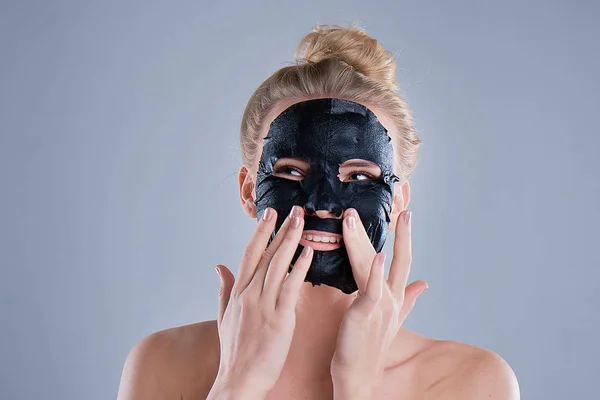 Menina com ombros nus posando em fundo cinza com máscara facial preta, conceito de cuidados com a pele, foto de beleza, tratamento de pele, máscara de pele hidratante, sorrindo . — Fotografia de Stock