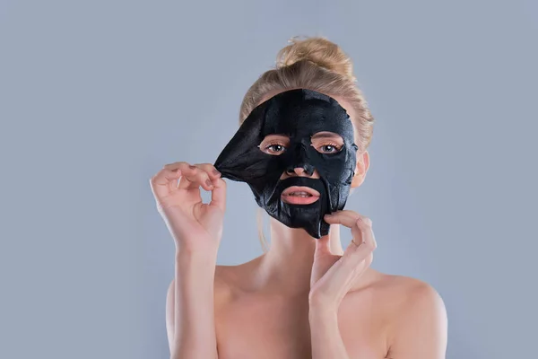 Flicka med nakna axlar poserar på grå bakgrund med ansiktsmask svart, hudvård koncept, skönhet foto, hudbehandling, återfuktande hud mask, leende. — Stockfoto