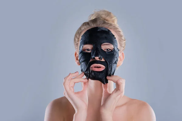 Девушка с обнаженными плечами позирует на сером фоне с черной маской для лица, концепция ухода за кожей, красота фото, лечение кожи, увлажняющая маска кожи, улыбаясь . — стоковое фото