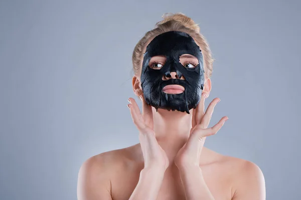 Ragazza con spalle nude in posa su sfondo grigio con maschera viso nero, concetto di cura della pelle, foto di bellezza, trattamento della pelle, idratante maschera della pelle, sorridente . — Foto Stock