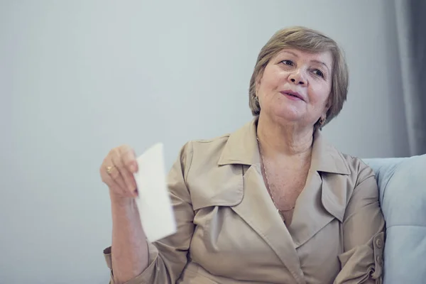 Mujer anciana sobrecalentada sentada en el sofá agitando ventilador para refrescarse, sesenta años mujer se siente mal caliente, cambios hormonales de edad, apartamentos sin aire acondicionado, concepto de malestar veraniego —  Fotos de Stock