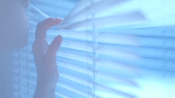 Intrigued pretty young woman looks out the window through the blinds then closes them by passing them by her hand. Close up of a hand reaching and opening a crack in the window blinds — Stock Video