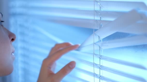 Intrigued pretty young woman looks out the window through the blinds then closes them by passing them by her hand. Close up of a hand reaching and opening a crack in the window blinds — Stock Video