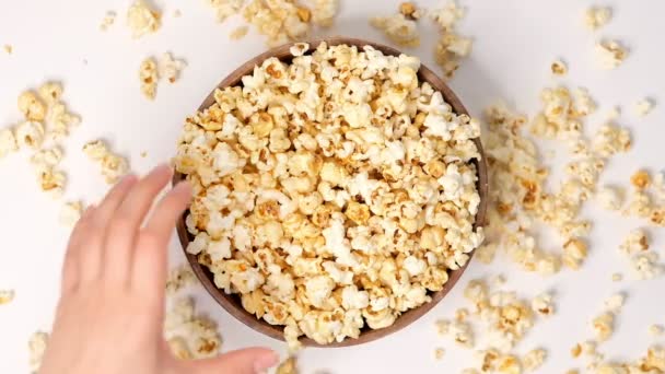 Palomitas de maíz arrojadas en un tazón de madera sobre un fondo blanco. Vídeo en cámara lenta. La mano femenina toma palomitas. Vista superior cercana — Vídeos de Stock