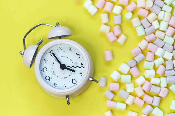 Vista dall'alto di Pastello a forma di Marshmallow caramelle e sveglia con alcuni sparsi sul pallido gialloTabella — Foto Stock