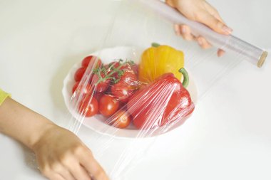 Beyaz bir masada yemek depolamak için yemek filmi kullanan bir kadın. Ürünleri paketlemek için şeffaf polietilen gıda filmi rulosu.