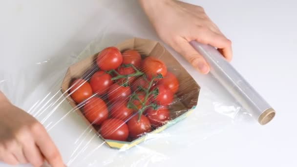 Kvinna som använder matfilm för förvaring av mat på ett vitt bord. Rulla av transparent polyeten livsmedelsfilm för förpackningsprodukter. körsbärstomater i engångsförpackningar av plast Slow Motion video. Närbild — Stockvideo