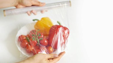 Beyaz bir masada yemek depolamak için yemek filmi kullanan bir kadın. Ürünleri paketlemek için şeffaf polietilen gıda filmi rulosu.