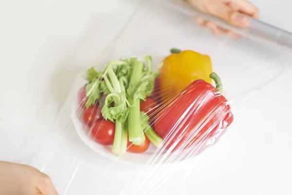 Beyaz bir masada yemek depolamak için yemek filmi kullanan bir kadın. Ürünleri paketlemek için şeffaf polietilen gıda filmi rulosu.