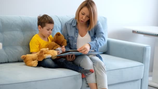 Mam en zoon lezen een boek zittend op een turquoise bank in de woonkamer. — Stockvideo