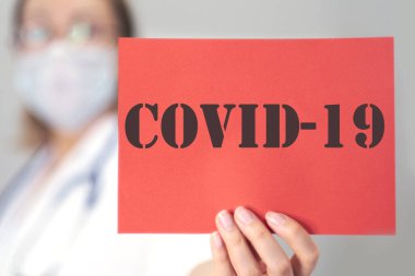 Doktor elinde COVID-19 yazılı kırmızı bir kart tutuyor. Mavi arkaplanlı Coronavirus 2019-NCoV romanından. Virüs Salgını Koruma Konsepti