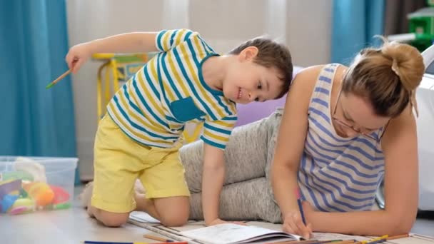 Jonge gelukkige moeder en zoontje liggen op de grond in de kinderkamer en doen kunsten en handwerk, tekenen samen — Stockvideo