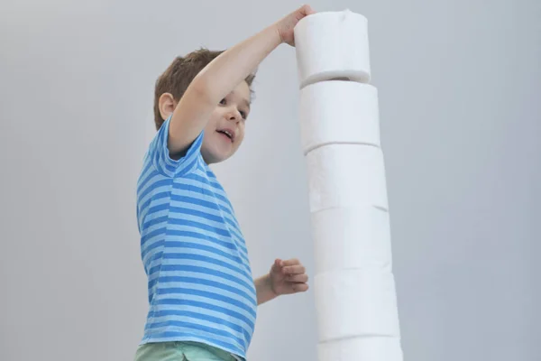 Pojken bygger ett torn av toalettpapper. Aktiv pojke leker med toalettpapper i retrofilter. Barnkramar en massa toalettpapper, Barnhälsovård koncept — Stockfoto