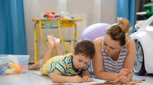 Νεαρή ευτυχισμένη μητέρα και ο μικρός γιος κείτονται στο πάτωμα στο παιδικό δωμάτιο και κάνουν τέχνες και χειροτεχνίες, ζωγραφίζοντας μαζί — Φωτογραφία Αρχείου