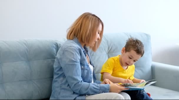 Мама и сын читают книгу, сидя на бирюзовом диване в гостиной . — стоковое видео