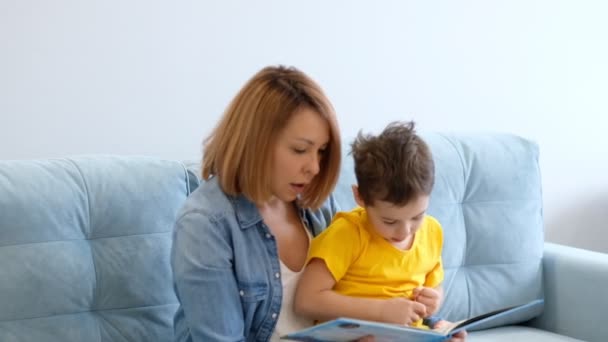 Anya és fia könyvet olvasnak egy türkiz kanapén ülve a nappaliban.. — Stock videók