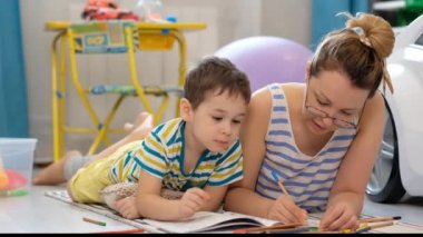 Genç mutlu anne ve küçük oğlu, çocuk odasında yerde uzanmış sanat ve el sanatları yapıyor, birlikte resim çiziyorlar.