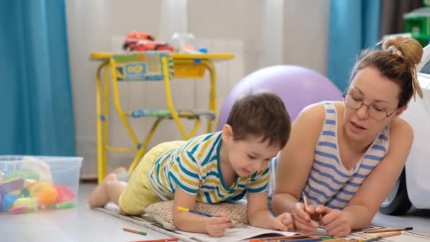 Jovem mãe feliz e pequeno filho deita-se no chão na sala das crianças e fazendo artes e ofícios, desenhando juntos — Vídeo de Stock