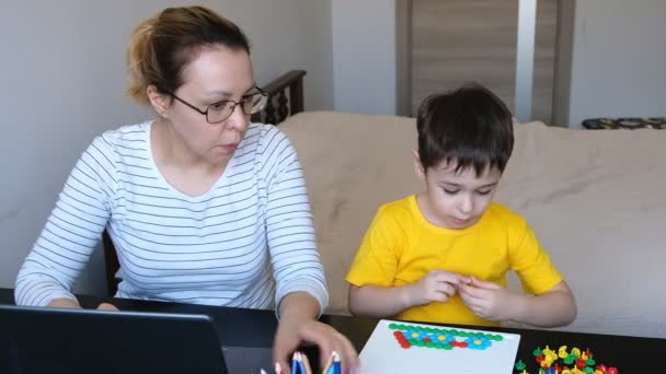 Mladá brunetka matka pracuje na svém notebooku se svým synem sedí na stole, hraje na svém tabletu. — Stock video