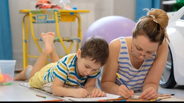 Молода щаслива мати і маленький син лежить на підлозі в дитячій кімнаті і займається мистецтвом і ремеслами, малюючи разом — стокове фото