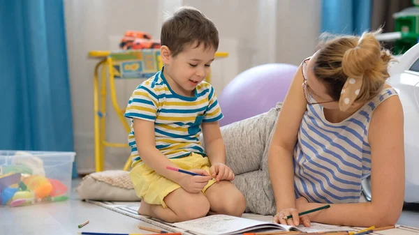 Молодая счастливая мать и маленький сын лежит на полу в детской комнате и занимается искусством и ремеслами, рисуя вместе — стоковое фото