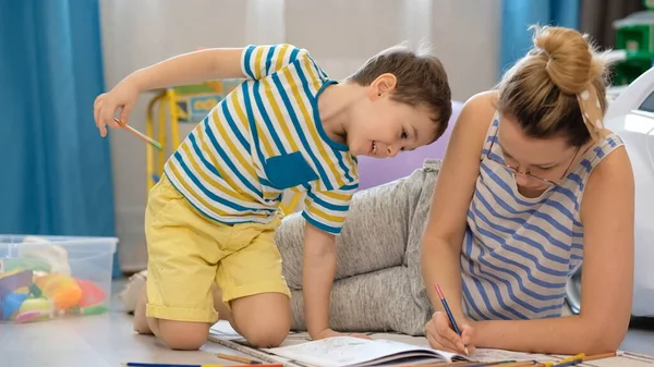 Young happy mother and little son lies on the floor in the childrens room and doing arts and crafts, drawing together — Stock Photo, Image