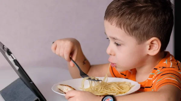 Drôle petit garçon manger des pâtes et regarder un dessin animé sur une tablette dans la table de cuisine — Photo