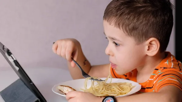 Śmieszne mały chłopiec jeść makaron i oglądać kreskówki na tablecie w kuchni tabeli — Zdjęcie stockowe