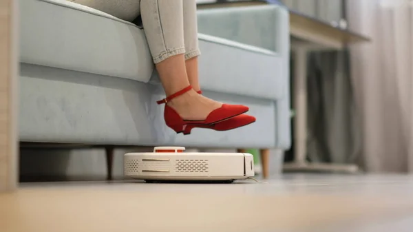 Aspiradora robótica limpiando la habitación mientras la mujer descansa en el sofá. — Foto de Stock