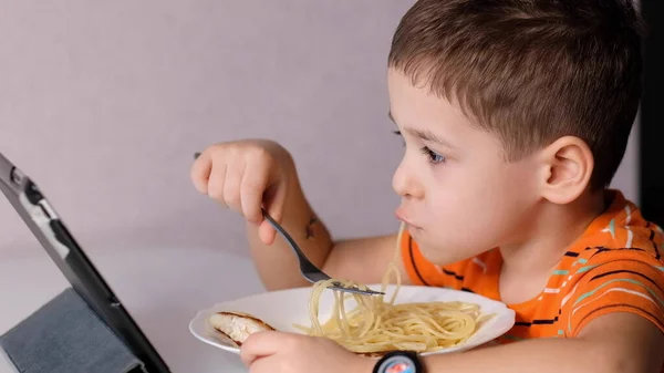 Engraçado menino comer macarrão e assistir a um desenho animado em um tablet na mesa da cozinha — Fotografia de Stock