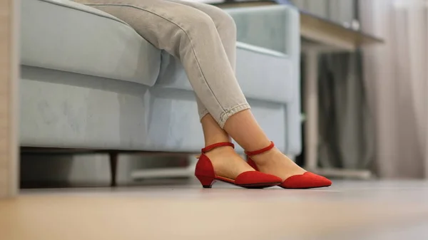 Mujer joven irreconocible en jeans ligeros descansando en el sofá. piernas cruzadas femeninas en zapatos rojos. casa, piso de madera gris —  Fotos de Stock