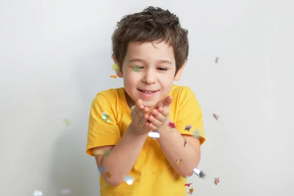 생일 축하 해. 매력적 이고 귀엽고 귀여운 작은 소년이 감정적 인 표정으로 축제 분위기를 보여 주기 위해 콘 페티를 불고 있는 사진. — 스톡 사진