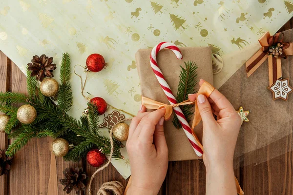 Vrouwen handen inpakken kerstcadeau, sluiten. Onvoorbereide kerstcadeaus op houten achtergrond met decor elementen en items, bovenaanzicht. Kerst of Nieuwjaar DIY verpakking Concept. — Stockfoto