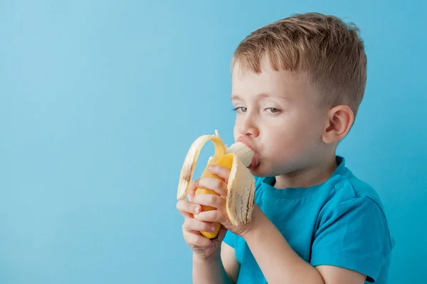 Little Boy Segurando e comendo uma banana no fundo azul, comida — Fotografia de Stock