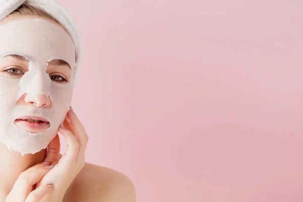 Belle jeune femme applique un masque de tissu cosmétique sur un visage sur fond rose — Photo