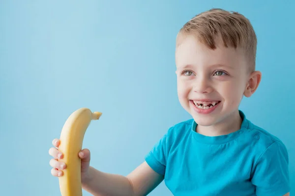 Niño sosteniendo y comiendo un plátano sobre fondo azul, comida — Foto de Stock