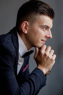 Endişeli damat elleriyle oynuyor ve smokin giyerken yan tarafa bakıyor, siyah stüdyo arka planında oturuyor.